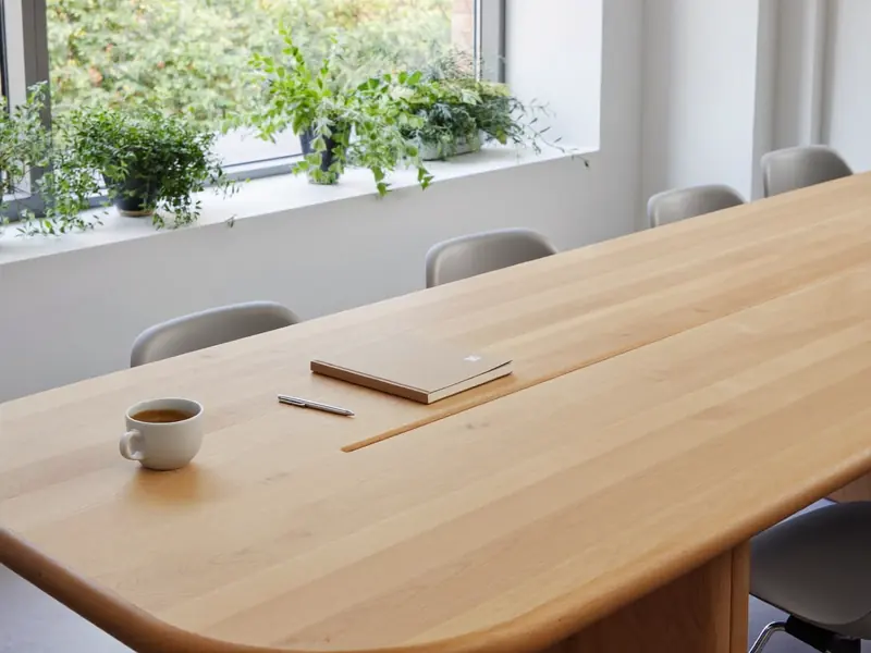 Heller Besprechungsraum mit Holztisch, Notizbuch, Kaffeetasse und Pflanzen am Fenster